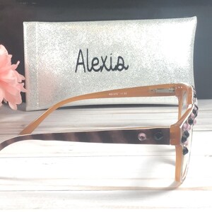 Op de afbeelding: Bruin gekleurde leesbril met decoratieve strass-steentjes op het montuur. De bril heeft een bruin en beige patroon op de poot. Een zilveren etui met de naam "Alexis" is op de achtergrond.