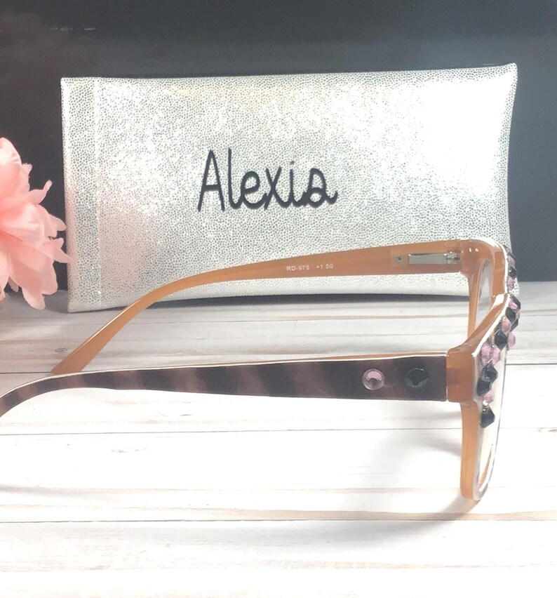 Op de afbeelding: Bruin gekleurde leesbril met decoratieve strass-steentjes op het montuur. De bril heeft een bruin en beige patroon op de poot. Een zilveren etui met de naam "Alexis" is op de achtergrond.