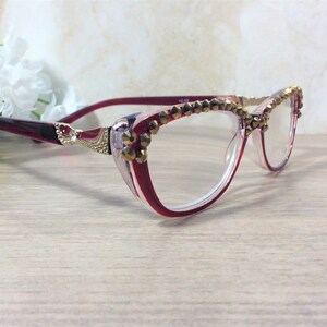 May include: Cat-eye reading glasses in red and clear with gold-coloured embellishments. The frames feature a decorative design at the temples and are adorned with small, gold-coloured studs along the top rim. The lenses are clear.