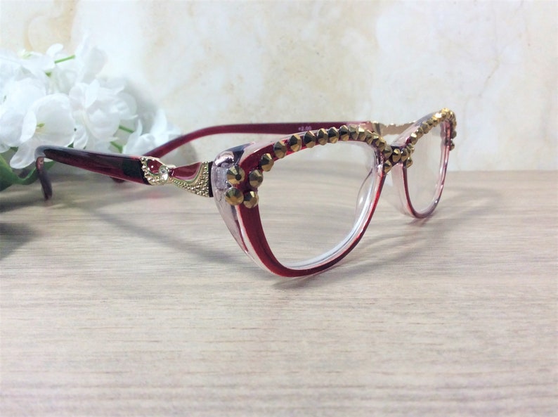May include: Cat-eye reading glasses in red and clear with gold-coloured embellishments. The frames feature a decorative design at the temples and are adorned with small, gold-coloured studs along the top rim. The lenses are clear.