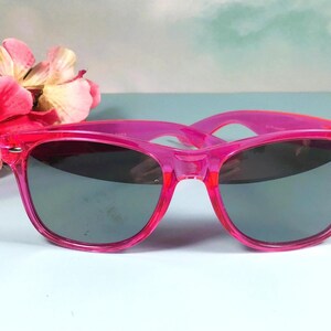 Op de afbeelding: Felroze zonnebril met donkere glazen. De monturen zijn doorschijnend, levendig roze. De zonnebril staat tegen een lichtblauwe achtergrond met roze bloemen. Een stijlvol accessoire.