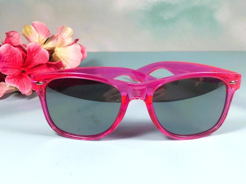 Op de afbeelding: Felroze zonnebril met donkere glazen. De monturen zijn doorschijnend, levendig roze. De zonnebril staat tegen een lichtblauwe achtergrond met roze bloemen. Een stijlvol accessoire.