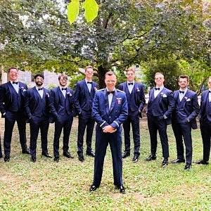 May include: A group of men in formal attire, including dark blue tuxedos with bow ties and black dress shoes, pose outdoors. The setting is a well-manicured lawn with a backdrop of lush green trees. The men are smiling and looking at the camera.