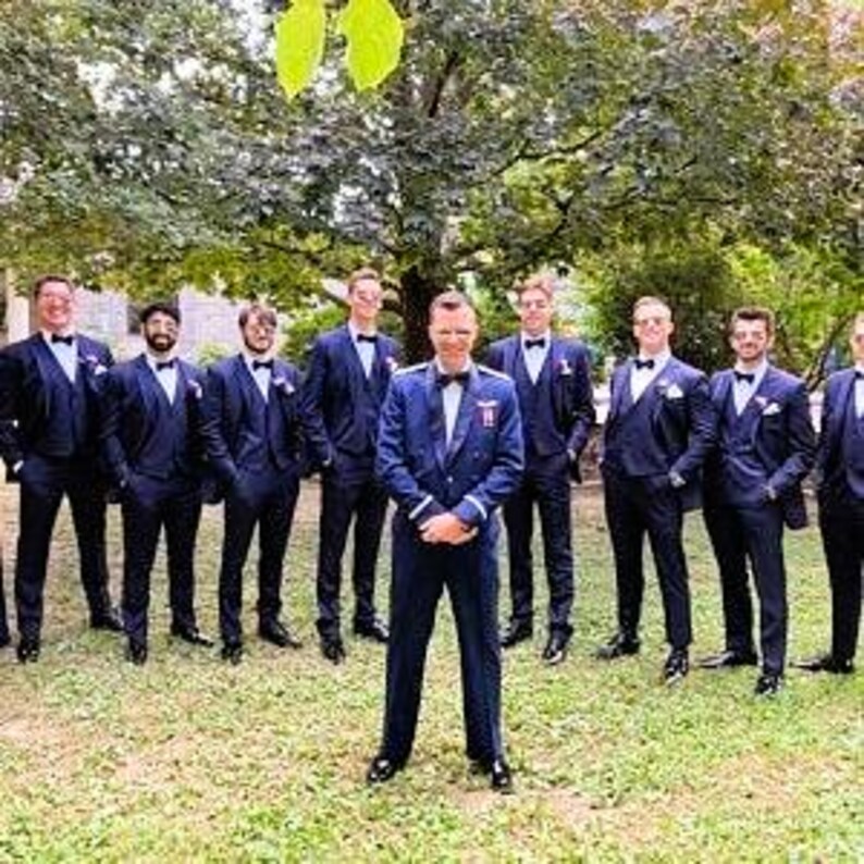 May include: A group of men in formal attire, including dark blue tuxedos with bow ties and black dress shoes, pose outdoors. The setting is a well-manicured lawn with a backdrop of lush green trees. The men are smiling and looking at the camera.