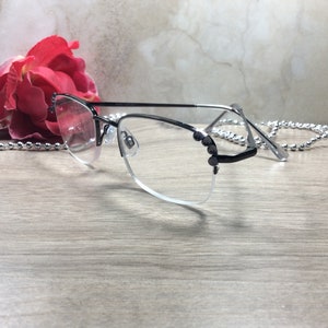 May include: A pair of eyeglasses with a black and silver frame, featuring a half-rim design. The glasses have clear lenses and are adorned with small, dark embellishments on the temples. A red rose and a silver beaded chain are in the background.