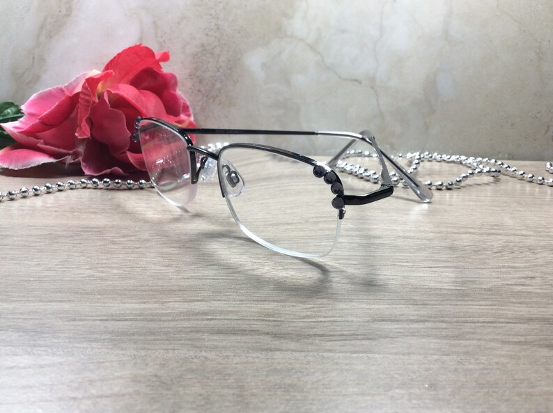May include: A pair of eyeglasses with a black and silver frame, featuring a half-rim design. The glasses have clear lenses and are adorned with small, dark embellishments on the temples. A red rose and a silver beaded chain are in the background.