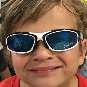Peut inclure: Un jeune gar&ccedil;on portant des lunettes de soleil noires et blanches avec des verres bleus. Les lunettes de soleil ont le nom "Hudson" inscrit sur la lentille droite.