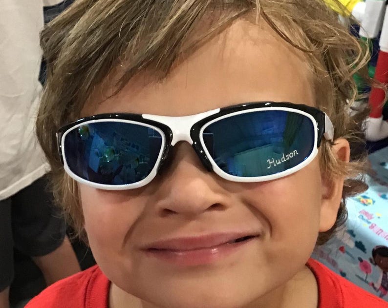 Peut inclure: Un jeune gar&ccedil;on portant des lunettes de soleil noires et blanches avec des verres bleus. Les lunettes de soleil ont le nom "Hudson" inscrit sur la lentille droite.