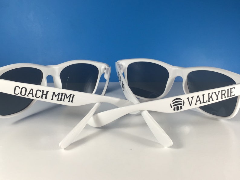 May include: Two pairs of white sunglasses with black lenses. One pair has the text "COACH MIMI" on the arm and the other pair has the text "VALKYRIE" and a volleyball icon on the arm.