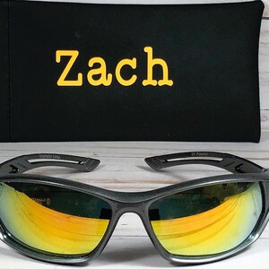 May include: Black sunglasses with yellow and orange lenses, resting in front of a black case. The case has the name "Zach" embroidered in yellow. The sunglasses have a sporty design.