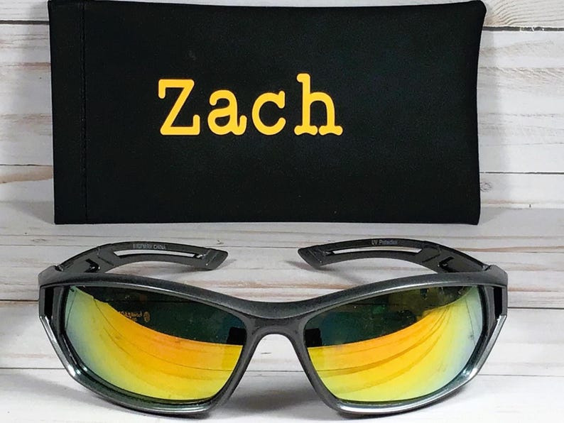 May include: Black sunglasses with yellow and orange lenses, resting in front of a black case. The case has the name "Zach" embroidered in yellow. The sunglasses have a sporty design.