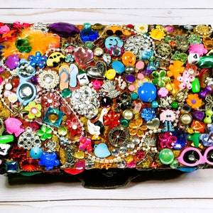 May include: A rectangular jewelry box covered in a mosaic of colorful embellishments. The box is adorned with various charms, including flowers, hearts, and beads. The color palette includes pink, blue, yellow, and silver, creating a vibrant and eclectic design.