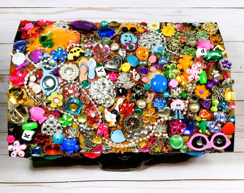 May include: A rectangular jewelry box covered in a mosaic of colorful embellishments. The box is adorned with various charms, including flowers, hearts, and beads. The color palette includes pink, blue, yellow, and silver, creating a vibrant and eclectic design.