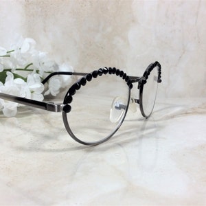 May include: Oval-shaped eyeglasses with a dark gray metal frame and clear lenses. The upper rims are adorned with a row of small, black decorative beads. The glasses are positioned in front of white flowers.