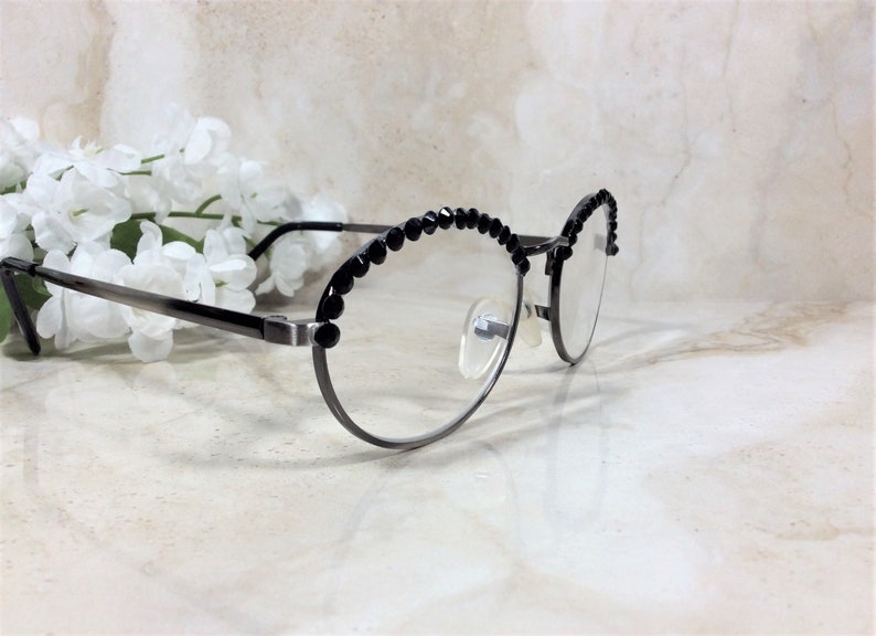 May include: Oval-shaped eyeglasses with a dark gray metal frame and clear lenses. The upper rims are adorned with a row of small, black decorative beads. The glasses are positioned in front of white flowers.
