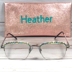 May include: A pair of eyeglasses with silver frames and iridescent crystals along the top rims, positioned in front of a rectangular rose gold glitter case featuring the name Heather printed in teal letters on the front surface.