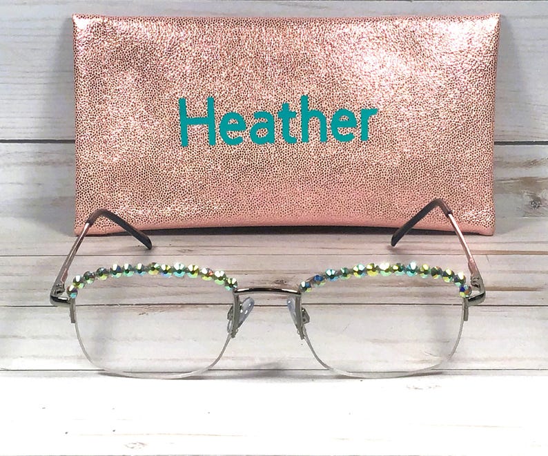 May include: A pair of eyeglasses with silver frames and iridescent crystals along the top rims, positioned in front of a rectangular rose gold glitter case featuring the name Heather printed in teal letters on the front surface.