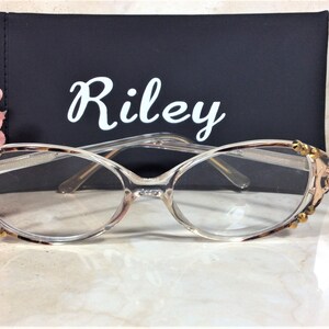 Puede incluir: Gafas con montura transparente con un patr&oacute;n de carey en los bordes superiores y las patillas, adornadas con adornos dorados. Las gafas est&aacute;n delante de un estuche negro con el nombre "Riley" en escritura blanca. Una flor rosa est&aacute; en el fondo.