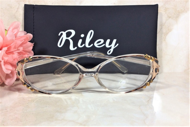 Puede incluir: Gafas con montura transparente con un patr&oacute;n de carey en los bordes superiores y las patillas, adornadas con adornos dorados. Las gafas est&aacute;n delante de un estuche negro con el nombre "Riley" en escritura blanca. Una flor rosa est&aacute; en el fondo.
