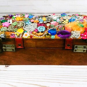May include: A wooden treasure chest with a dark brown finish, adorned with an array of colorful embellishments. The top is covered in various jewels, beads, and trinkets, including a smiley face, flowers, and other decorative items. The chest has metal hinges and a red leather strap.