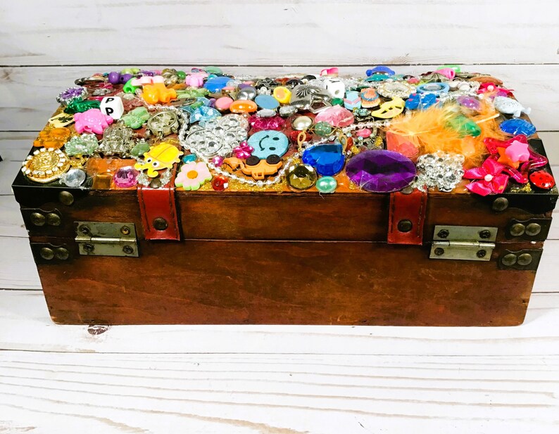 May include: A wooden treasure chest with a dark brown finish, adorned with an array of colorful embellishments. The top is covered in various jewels, beads, and trinkets, including a smiley face, flowers, and other decorative items. The chest has metal hinges and a red leather strap.