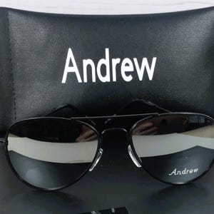 May include: Black aviator sunglasses with a thin metal frame and reflective lenses. The right lens features the white text Andrew. The sunglasses are positioned in front of a black rectangular case with the white text Andrew printed on the front.