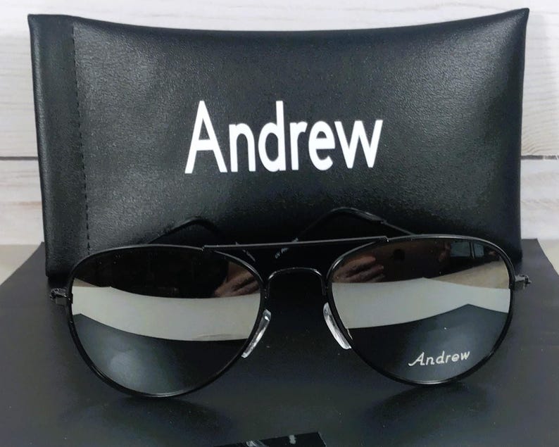 May include: Black aviator sunglasses with a thin metal frame and reflective lenses. The right lens features the white text Andrew. The sunglasses are positioned in front of a black rectangular case with the white text Andrew printed on the front.