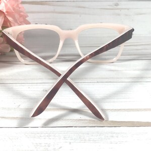 May include: Pale pink eyeglasses with brown tortoiseshell arms. The glasses have a square frame and are set against a white wooden background with pink flowers. The arms are crossed.