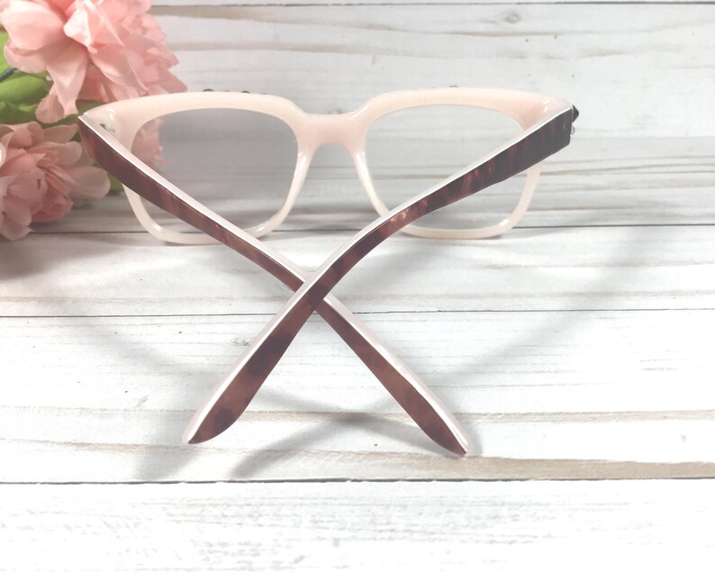 May include: Pale pink eyeglasses with brown tortoiseshell arms. The glasses have a square frame and are set against a white wooden background with pink flowers. The arms are crossed.