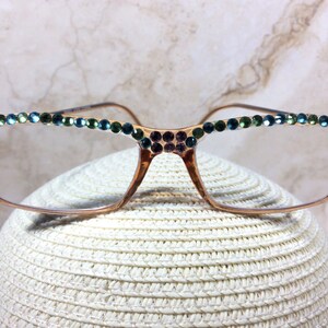 May include: Gold-toned eyeglasses with rectangular frames, adorned with rows of colorful rhinestones. The top of the frames and the temples are embellished with small, sparkling gems in shades of green, blue, and purple. The glasses are resting on a white hat.