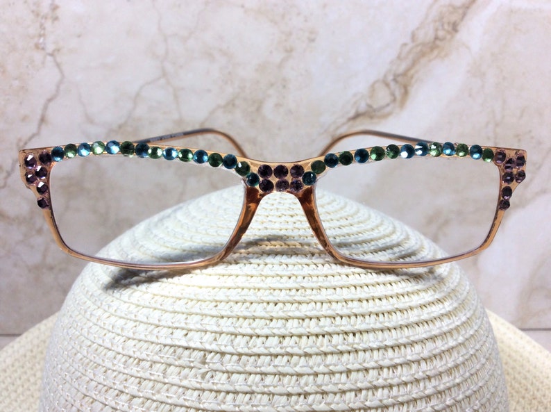 May include: Gold-toned eyeglasses with rectangular frames, adorned with rows of colorful rhinestones. The top of the frames and the temples are embellished with small, sparkling gems in shades of green, blue, and purple. The glasses are resting on a white hat.