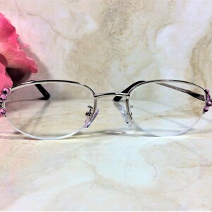 May include: Silver-rimmed spectacles with clear lenses. The frames feature a half-rim design and are adorned with small, pink gemstones at the temples. The glasses are set against a soft, neutral background, with a pink flower in the upper left corner.