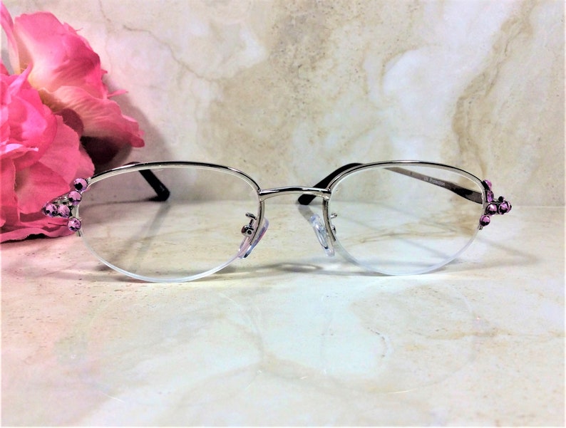May include: Silver-rimmed spectacles with clear lenses. The frames feature a half-rim design and are adorned with small, pink gemstones at the temples. The glasses are set against a soft, neutral background, with a pink flower in the upper left corner.