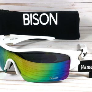 May include: White sports sunglasses with a rainbow iridescent lens. The sunglasses have a black case with the word "BISON" printed on it. A white sign says "Name Engraved FREE".
