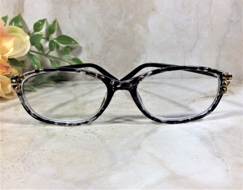 Peut inclure: Lunettes avec une monture au motif mouchet&eacute; noir et blanc. Les coins ext&eacute;rieurs de la monture sont orn&eacute;s de petits clous m&eacute;talliques dor&eacute;s. Les branches sont enti&egrave;rement noires et les verres sont clairs et transparents.