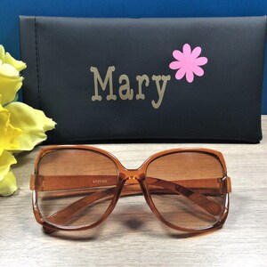 May include: Brown-framed sunglasses with tinted lenses. A black case with the name "Mary" and a pink flower graphic is behind the sunglasses. The sunglasses have a retro, oversized design.