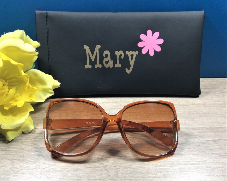 May include: Brown-framed sunglasses with tinted lenses. A black case with the name "Mary" and a pink flower graphic is behind the sunglasses. The sunglasses have a retro, oversized design.
