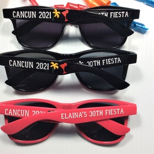Op de afbeelding: Drie zonnebrillen met zwarte monturen en donkere glazen. De bovenste twee paren zijn zwart met witte tekst die "CANCUN 2021" en "30TH FIESTA" zegt. Het onderste paar is roze met witte tekst die "CANCUN 2021" en "ELAINAS'S 30TH FIESTA" zegt.