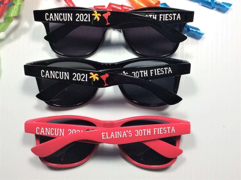 Op de afbeelding: Drie zonnebrillen met zwarte monturen en donkere glazen. De bovenste twee paren zijn zwart met witte tekst die "CANCUN 2021" en "30TH FIESTA" zegt. Het onderste paar is roze met witte tekst die "CANCUN 2021" en "ELAINAS'S 30TH FIESTA" zegt.
