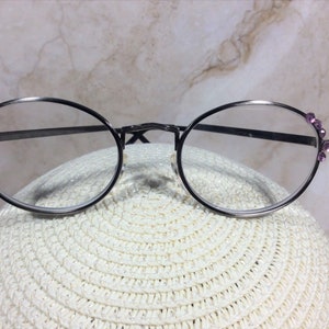 Puede incluir: Gafas redondas con montura de metal oscuro y lentes transparentes. Las patillas est&aacute;n adornadas con peque&ntilde;as gemas moradas. Las gafas descansan sobre una superficie blanca y texturizada.