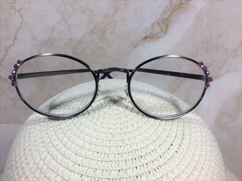 Puede incluir: Gafas redondas con montura de metal oscuro y lentes transparentes. Las patillas est&aacute;n adornadas con peque&ntilde;as gemas moradas. Las gafas descansan sobre una superficie blanca y texturizada.