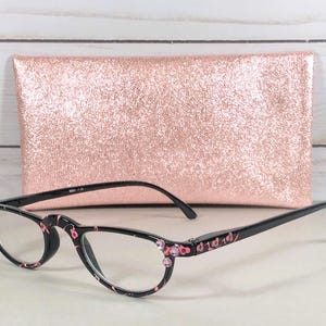 Pu&ograve; includere: Occhiali da lettura neri con un motivo floreale rosa sulle montature, accentuati da piccoli strass rosa agli angoli esterni. Gli occhiali sono abbinati a una custodia rettangolare con una texture glitterata color oro rosa.