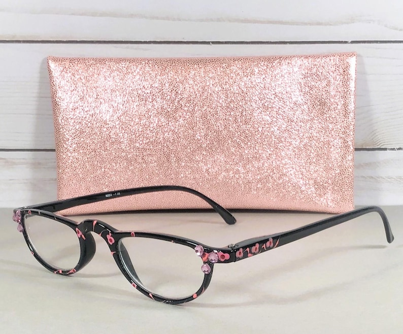 Pu&ograve; includere: Occhiali da lettura neri con un motivo floreale rosa sulle montature, accentuati da piccoli strass rosa agli angoli esterni. Gli occhiali sono abbinati a una custodia rettangolare con una texture glitterata color oro rosa.