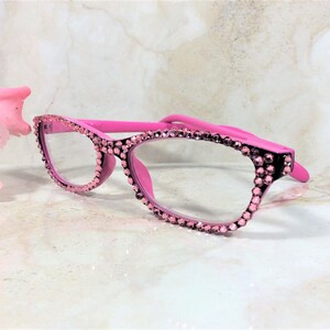 Pu&ograve; includere: Occhiali rosa con finiture di strass. La montatura &egrave; di un rosa acceso e gli strass sono di un rosa pi&ugrave; chiaro. Gli occhiali sono su uno sfondo morbido e sfocato con un fiore rosa.