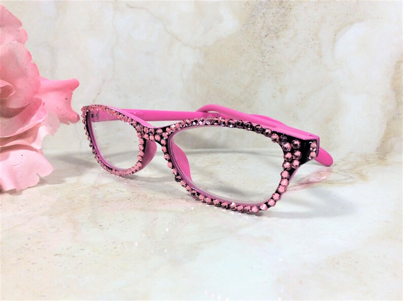 Pu&ograve; includere: Occhiali rosa con finiture di strass. La montatura &egrave; di un rosa acceso e gli strass sono di un rosa pi&ugrave; chiaro. Gli occhiali sono su uno sfondo morbido e sfocato con un fiore rosa.