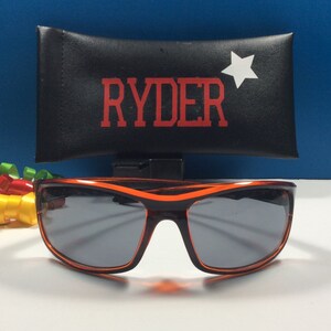 K&ouml;nnte beinhalten: Orangefarbene Sonnenbrille mit schwarzem Rahmen und grauen Gl&auml;sern. Die Sonnenbrille befindet sich in einem schwarzen Etui mit dem Namen "RYDER" und einem wei&szlig;en Stern, der darauf gedruckt ist.