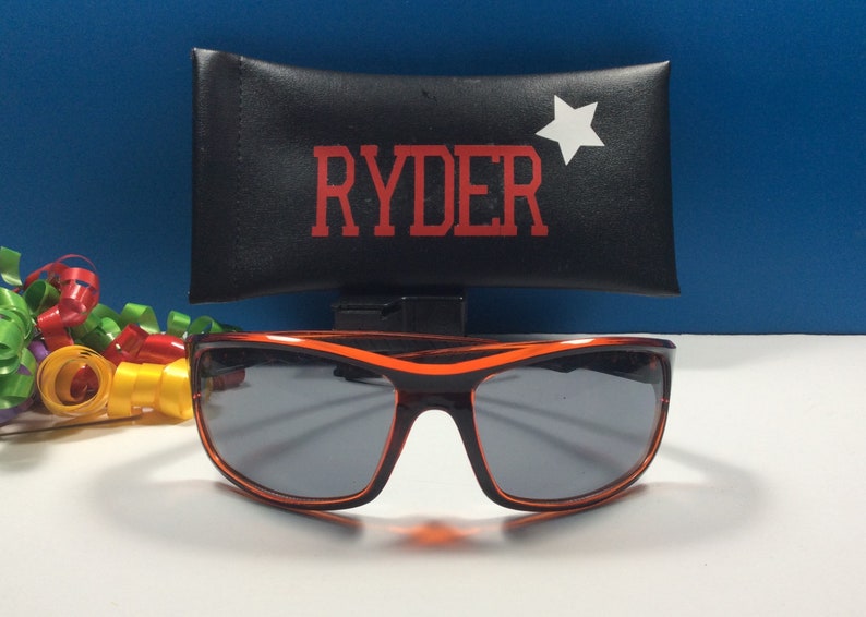 K&ouml;nnte beinhalten: Orangefarbene Sonnenbrille mit schwarzem Rahmen und grauen Gl&auml;sern. Die Sonnenbrille befindet sich in einem schwarzen Etui mit dem Namen "RYDER" und einem wei&szlig;en Stern, der darauf gedruckt ist.