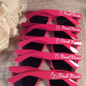 May include: Five pairs of pink plastic sunglasses with black lenses. Each pair has the text "Final Fiesta" and a small graphic of either a champagne flute or a wedding ring.