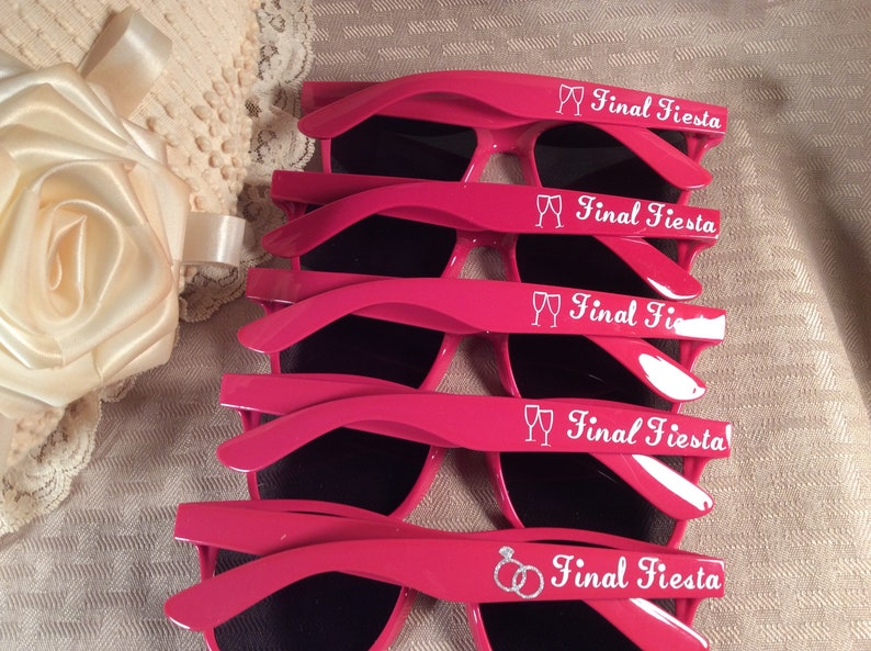 May include: Five pairs of pink plastic sunglasses with black lenses. Each pair has the text "Final Fiesta" and a small graphic of either a champagne flute or a wedding ring.