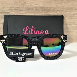 Puede incluir: Gafas de sol negras con lentes iridiscentes y detalles de estrellas plateadas. Las gafas tienen el nombre "Liliana" grabado en la lente derecha y una etiqueta que dice "Nombre grabado GRATIS".